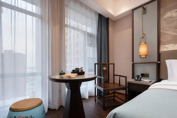 A hotel room with a bed, a desk with a telephone, a chair, a small table with books, and a window with sheer curtains.