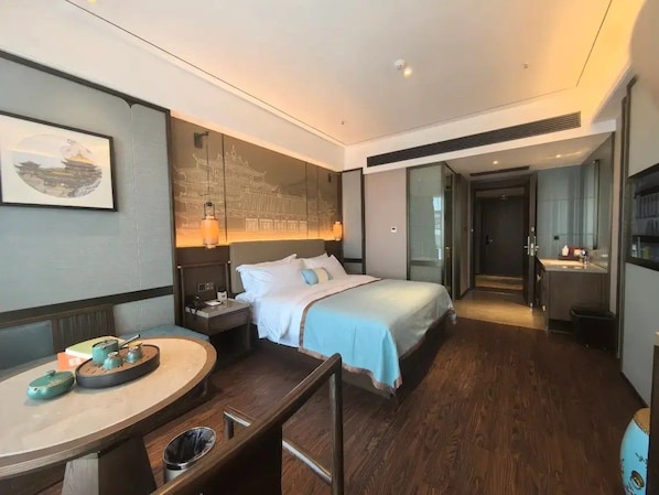 A hotel room with a bed, bedside table, and a desk with a chair. There is a wall-mounted artwork and a window.