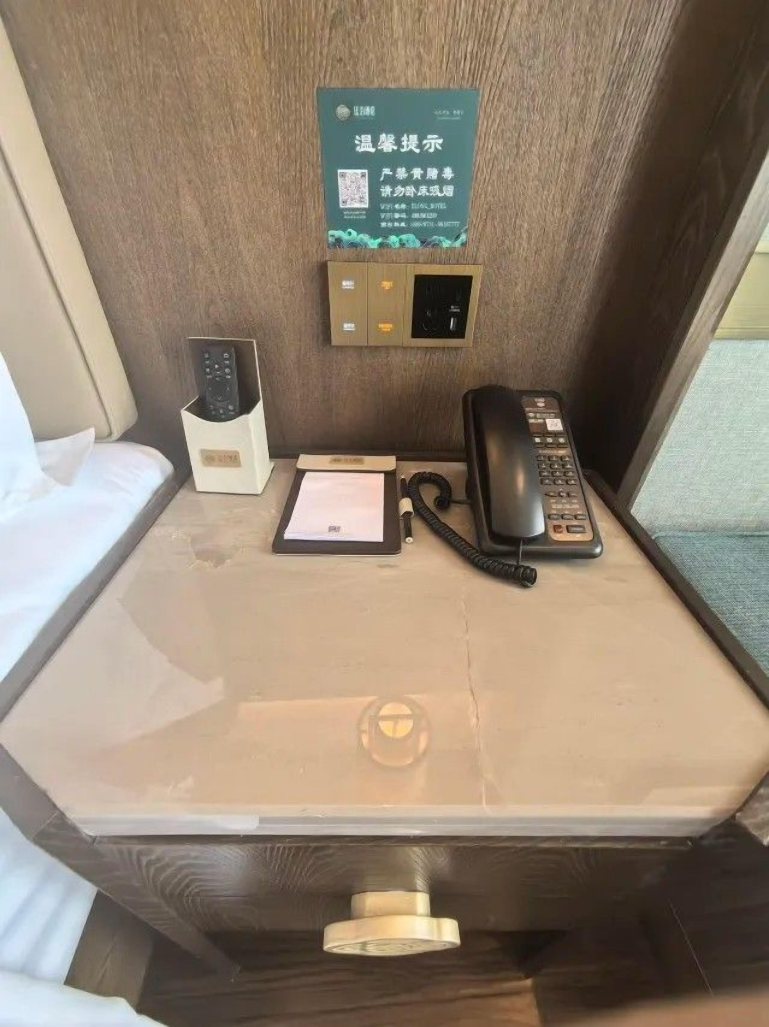 Habitación de hotel con mesita de noche, teléfono, control remoto y un cartel con código QR.