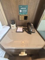 Habitación de hotel con mesita de noche, teléfono, control remoto y un cartel con código QR.