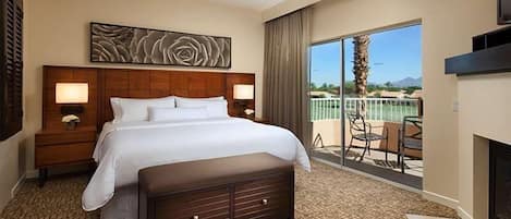 A hotel room with a large bed, a bench, a nightstand, a wall-mounted artwork, a fireplace, and a balcony with a view.