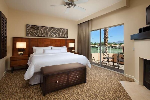 A hotel room with a large bed, a bench, a nightstand, a wall-mounted artwork, a fireplace, and a balcony with a view.