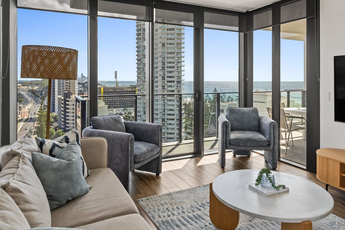 A modern living room with a sofa, armchairs, a coffee table, and a view of the city and ocean through large windows.