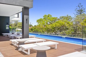 A poolside area with white lounge chairs and tables, a clear blue pool, and a view of trees and buildings in the background. - City Views, Ocean Hues | Sandbar (Gold Coast)
