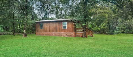 A wooden cabin with a deck and a staircase, surrounded by a grassy field and trees.