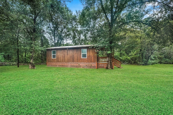 A wooden cabin with a deck and a staircase, surrounded by a grassy field and trees.