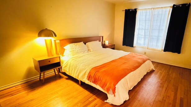 Un dormitorio con piso de madera, una cama con ropa de cama blanca y naranja, una mesita de noche con una lámpara y una ventana con cortinas.