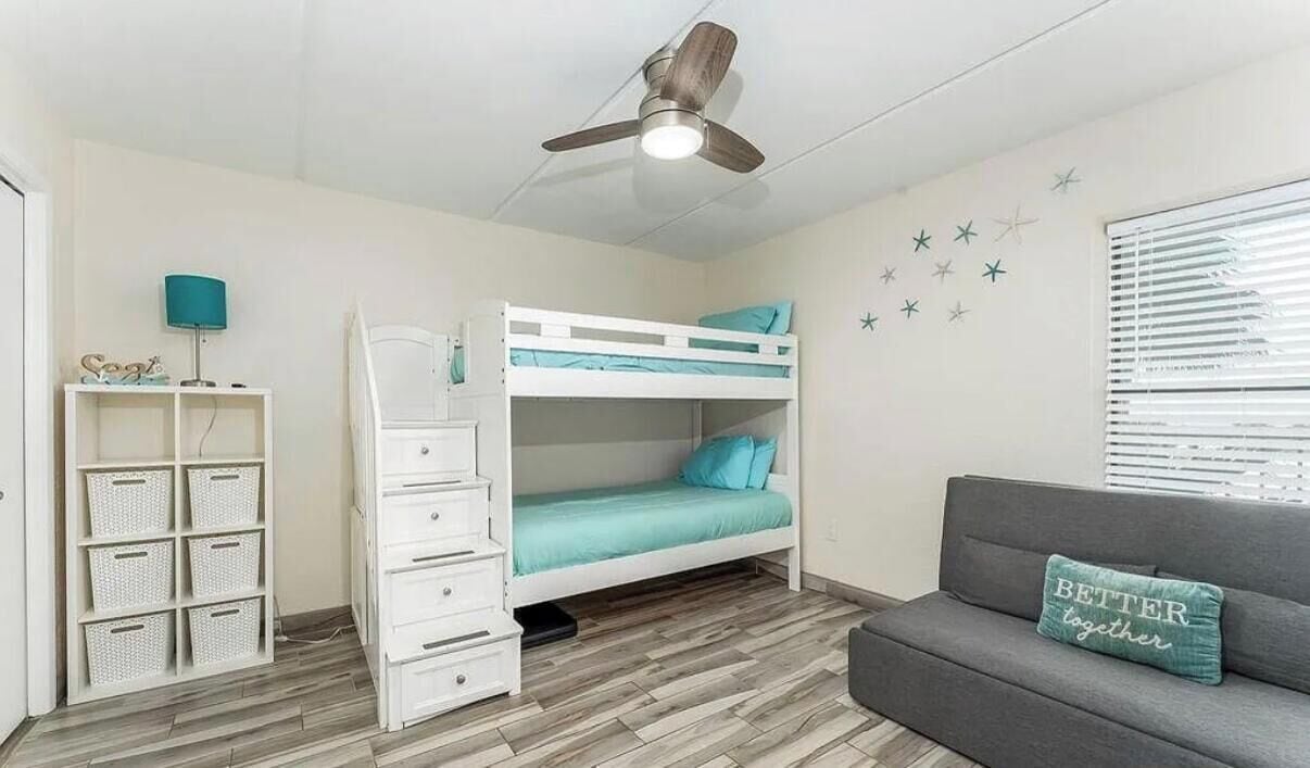 A room with a bunk bed, a desk with drawers, a grey sofa, a ceiling fan, and a window with blinds.