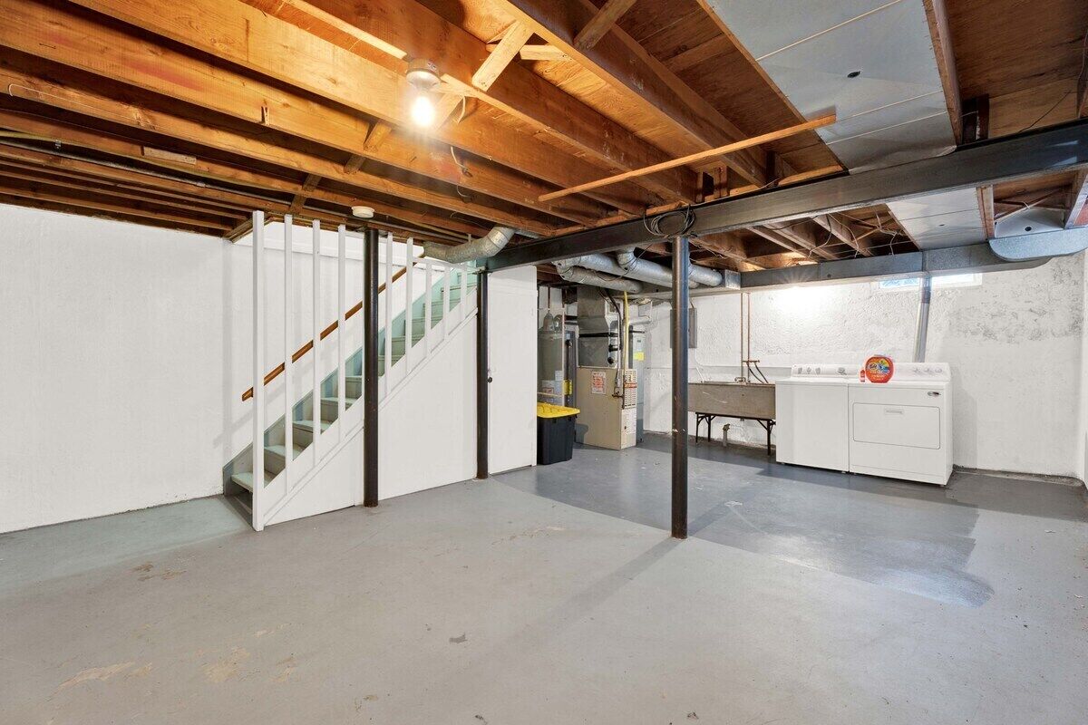 A basement with a concrete floor, wooden beams, a staircase, a washing machine, and a utility room with pipes.