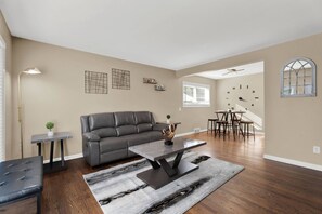 Ein geräumiges Wohnzimmer mit einem grauen Ledersofa, einem Couchtisch und einem Essbereich mit rundem Tisch und Stühlen.