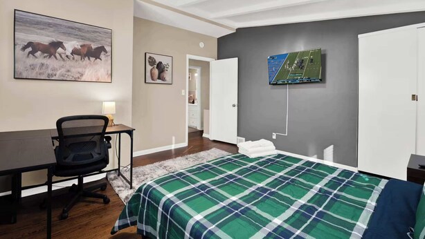 Quarto com cama, escrivaninha, cadeira, televisão na parede e porta que dá para outro cômodo.