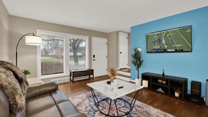 Uma sala de estar com sofá, mesa de centro, TV fixada na parede e um banco.
