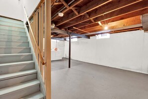 Un sótano con una escalera que sube, piso de hormigón y estructura de techo de madera.
