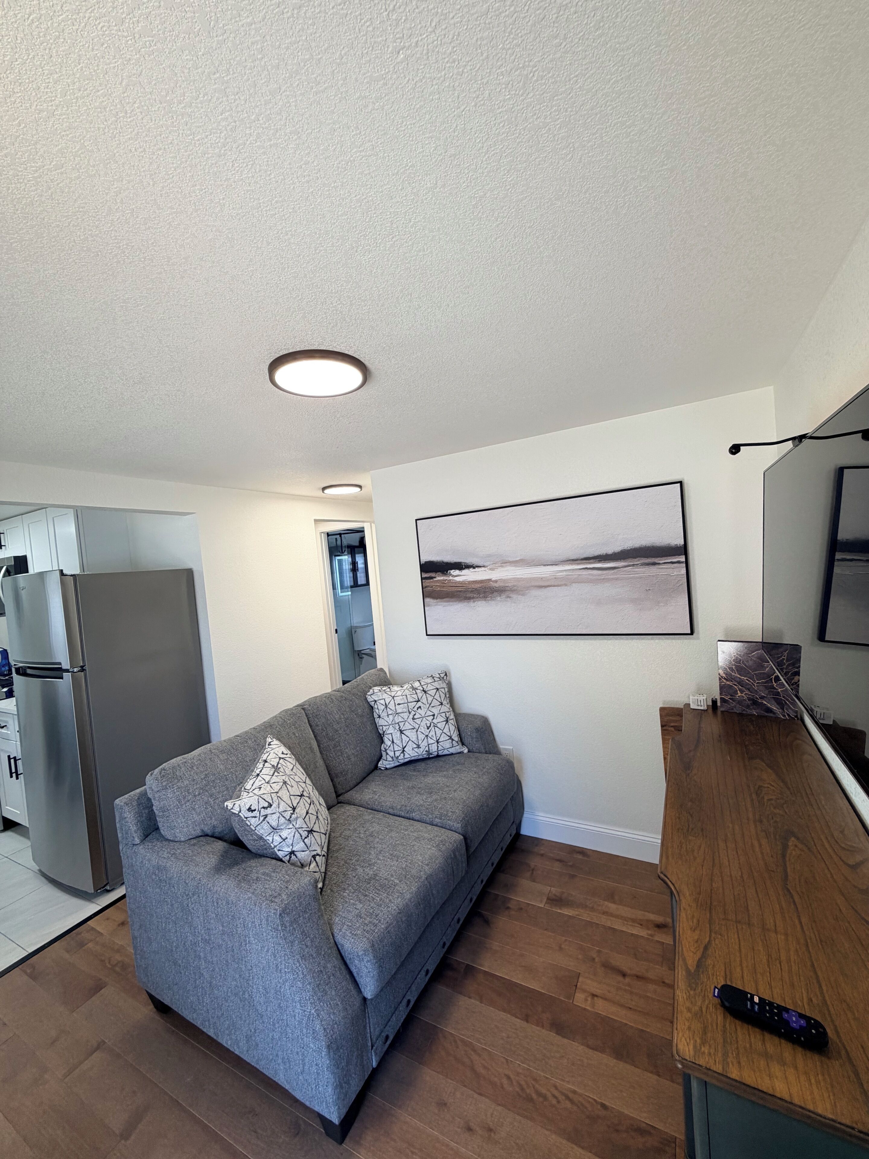 A compact living space with a grey sofa, a wooden coffee table, a flat-screen TV, and a refrigerator.