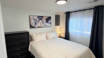 Una camera da letto con un letto, una cassettiera, un'opera d'arte appesa al muro, una finestra con tende e una lampada a soffitto.