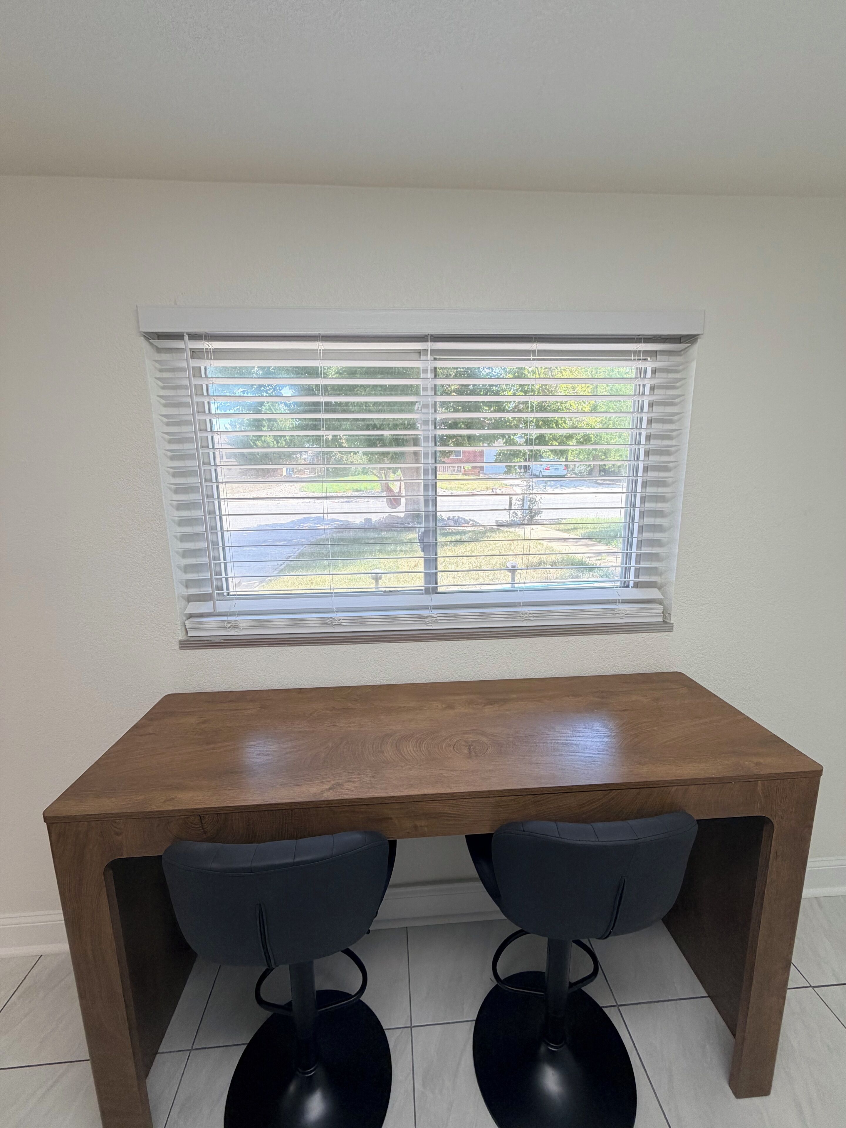 A room with a wooden desk, two black chairs, a window with blinds, and a view of a yard.