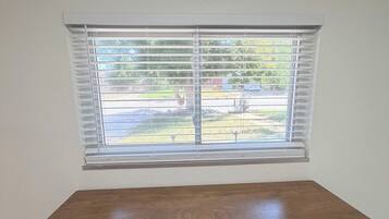 A room with a wooden desk, two black chairs, a window with blinds, and a view of a yard.