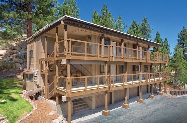 A multi-story wooden cabin with a balcony, surrounded by trees and a stone wall.