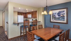 A compact hotel room with a kitchenette, a dining area with a wooden table and chairs, and a framed landscape painting on the wall.