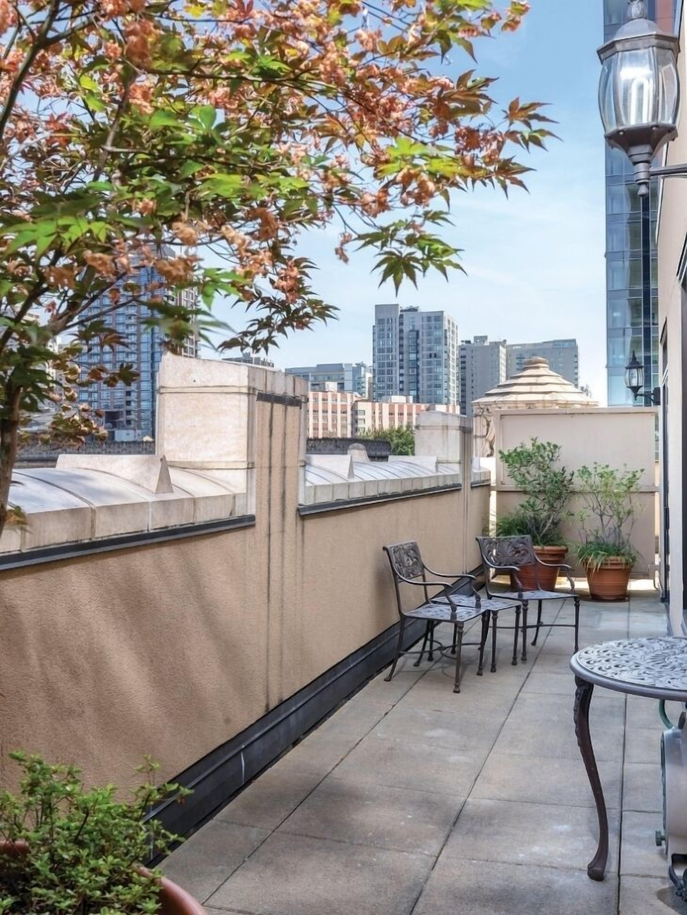 A rooftop terrace with a view of the city skyline, featuring a table and chairs, potted plants, and a lamp post.