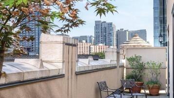 A rooftop terrace with a view of the city skyline, featuring a table and chairs, potted plants, and a lamp post.