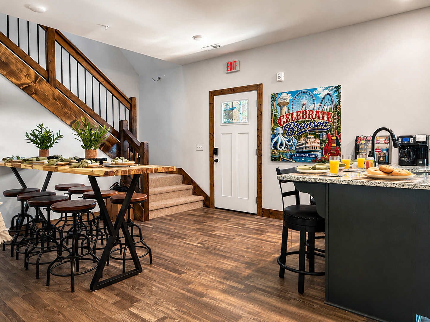 A kitchen with a wooden staircase, a dining area with stools, a coffee station, and a wall with a poster.