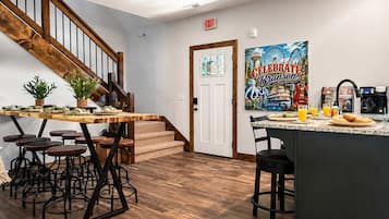 A kitchen with a wooden staircase, a dining area with stools, a coffee station, and a wall with a poster.