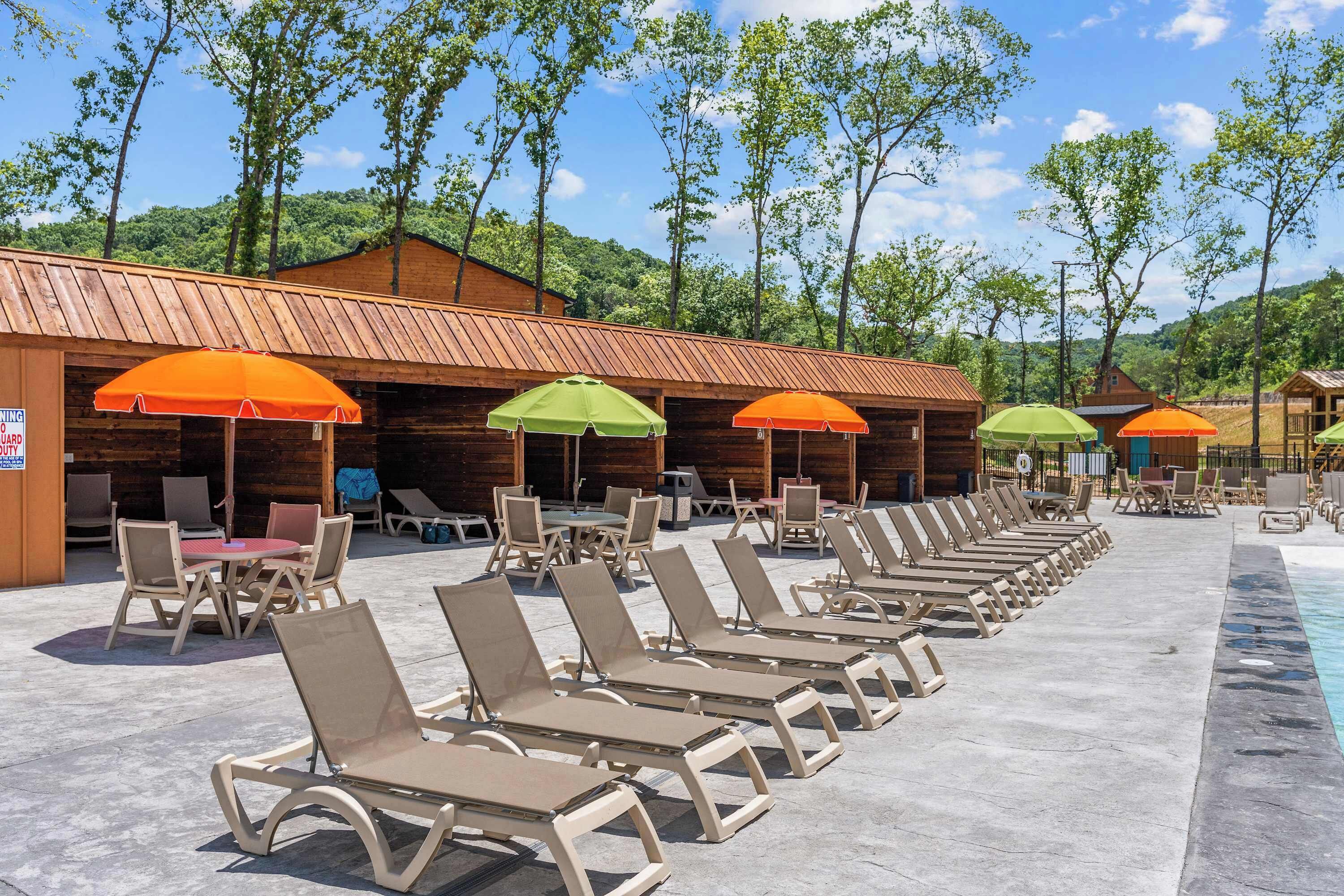 Zona junto a la piscina con sillones y sombrillas, cabañas de madera al fondo y cielo despejado.