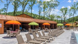 A poolside area with lounge chairs and umbrellas, wooden cabins in the background, and a clear sky.