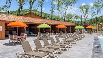 A poolside area with lounge chairs and umbrellas, wooden cabins in the background, and a clear sky.