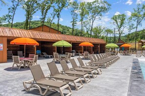 A poolside area with lounge chairs and umbrellas, wooden cabins in the background, and a clear sky.