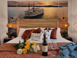 A bedroom with a large bed, a wall-mounted painting of a boat, two bedside tables with lamps, and a bouquet of flowers on the bed.