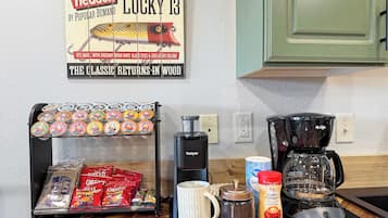 A kitchen counter with a coffee maker, a mug, a jar of coffee creamer, a kettle, a coffee grinder, and a snack rack with chips and cheese puffs.