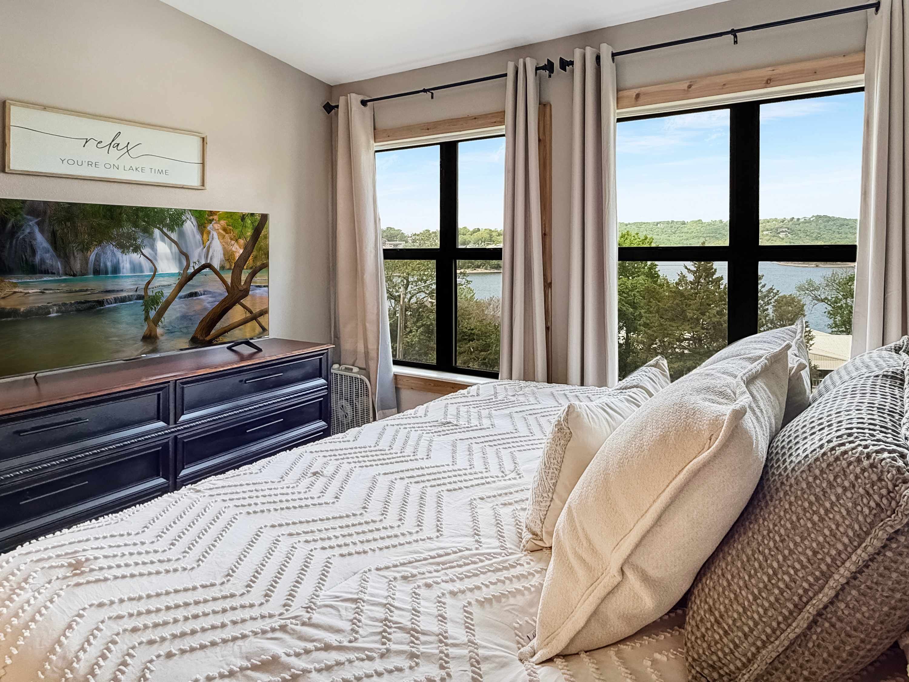 Ein Schlafzimmer mit Bett, Schminktisch, einem Gemälde und einem Fenster mit Blick auf einen See und Bäume.