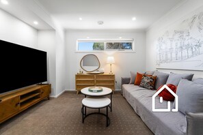A modern living room with a flat-screen TV, a round table, a sofa with cushions, a wooden dresser, and a wall-mounted mirror. - The Sorrento Sojourn *1 NIGHT FREE* Luxury Spa Retreat (Sorrento)