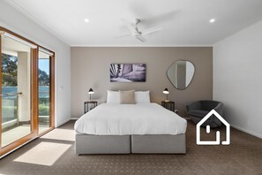 Ein Schlafzimmer mit einem Bett, zwei Nachttischen, einem Sessel, einem Deckenventilator, einem großen Fenster und einem wandmontierten Spiegel.