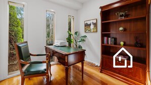 Una habitación con un escritorio de madera, una silla verde, una estantería y una ventana con vistas a la vegetación.