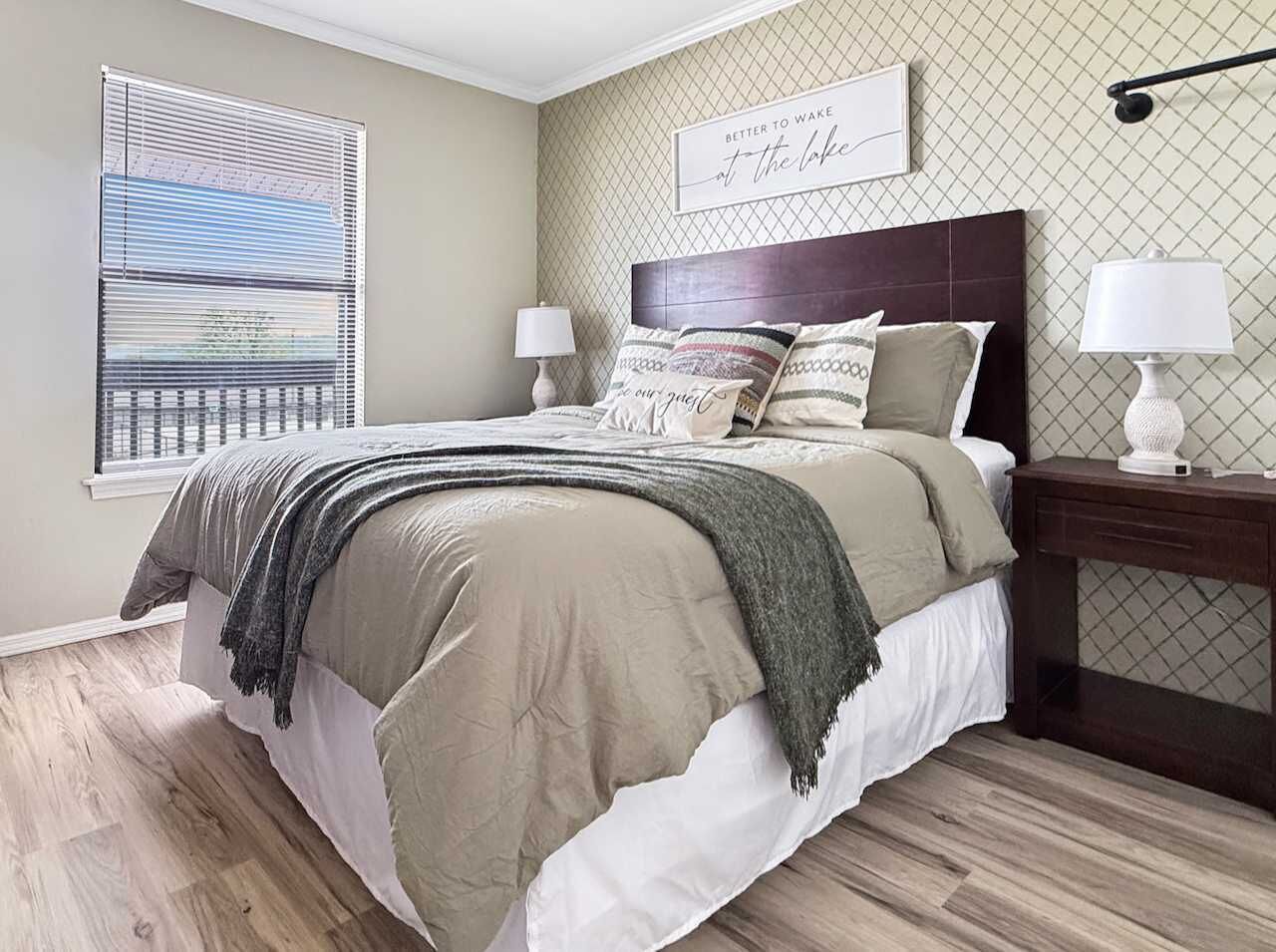 A bedroom with a bed, bedside tables, a window with blinds, and a wall-mounted lamp.