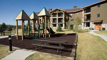 Un parque infantil con estructuras para escalar y una mesa de picnic, rodeado por un edificio con balcones y fachada de piedra.
