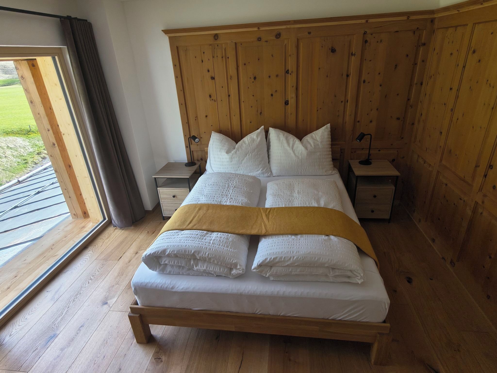 A bedroom with a wooden bed, two bedside tables, a window with curtains, and a view of the outdoors.