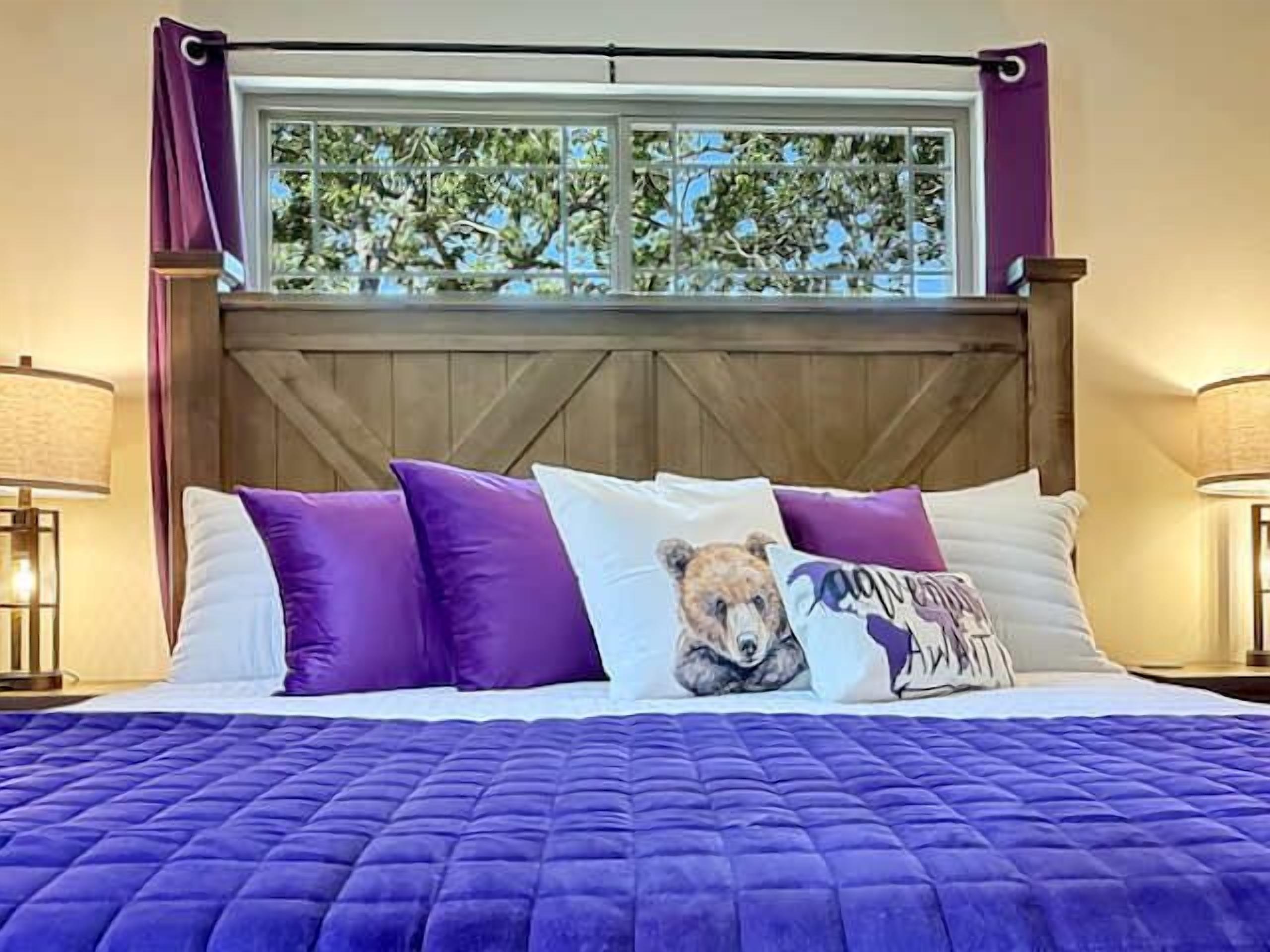Un letto con una coperta viola, una finestra vista alberi e una testiera in legno.