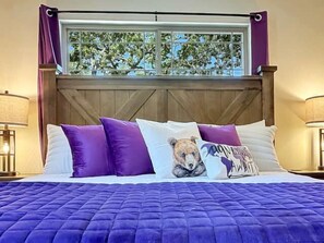 Una cama con colcha morada, ventana con vistas a árboles y cabecera de madera.
