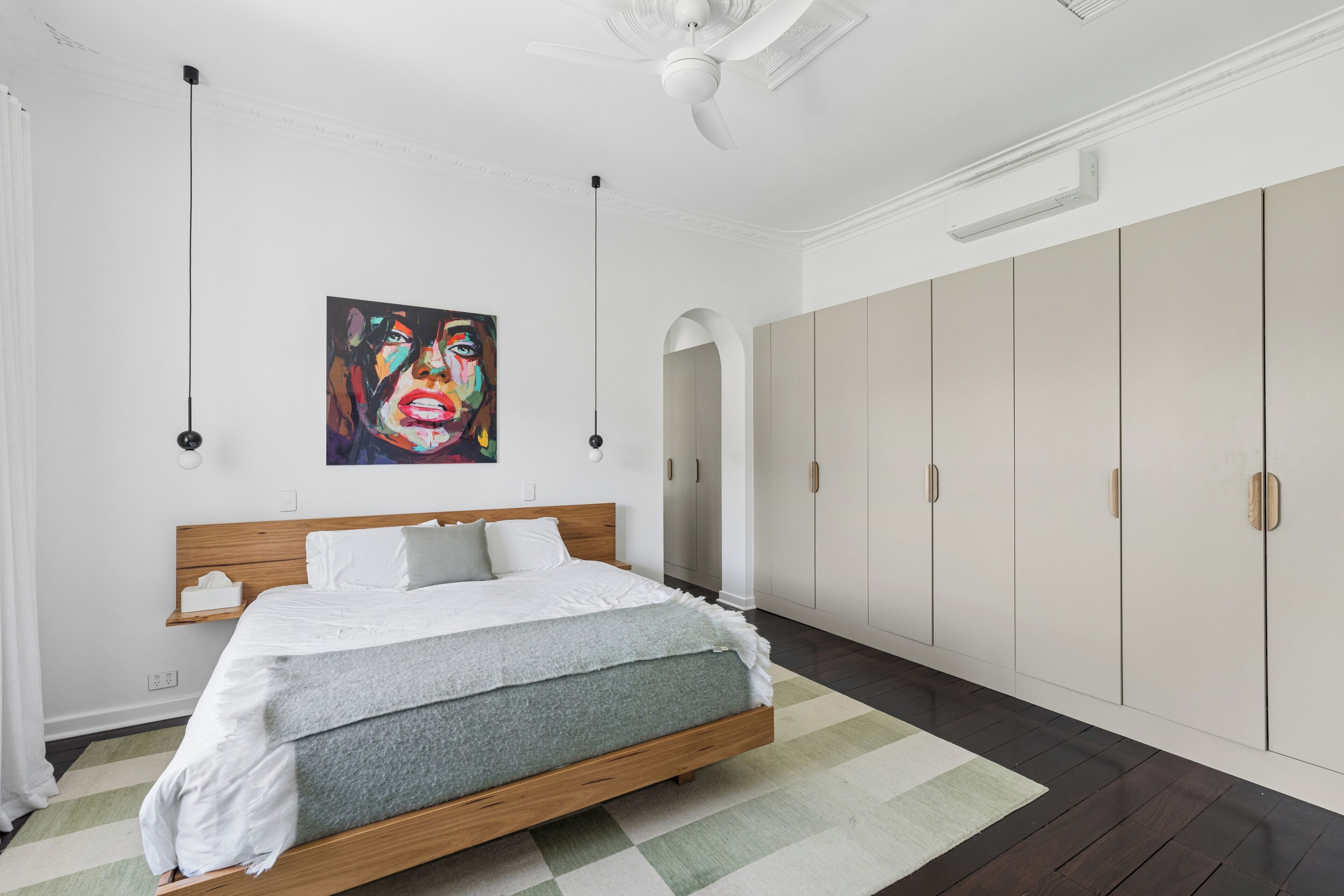 A bedroom with a bed, a large wardrobe, a ceiling fan, and a painting on the wall.