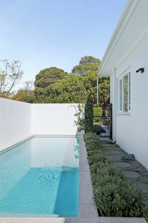 A swimming pool adjacent to a white building with a black gate, surrounded by greenery and trees.