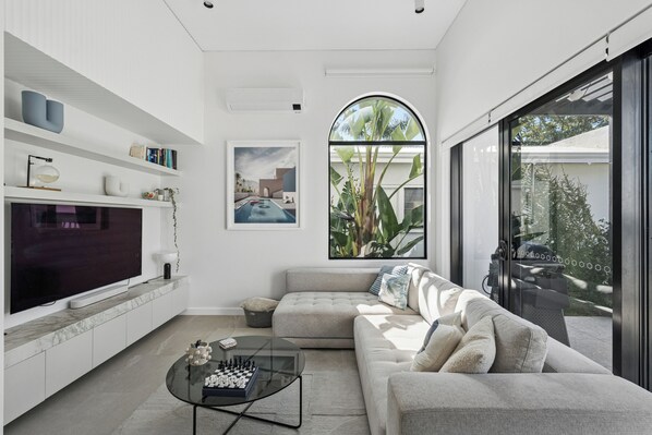A modern living room with a large flat-screen TV, a glass coffee table with a chess set, a grey sofa, and a large window with a view of greenery.