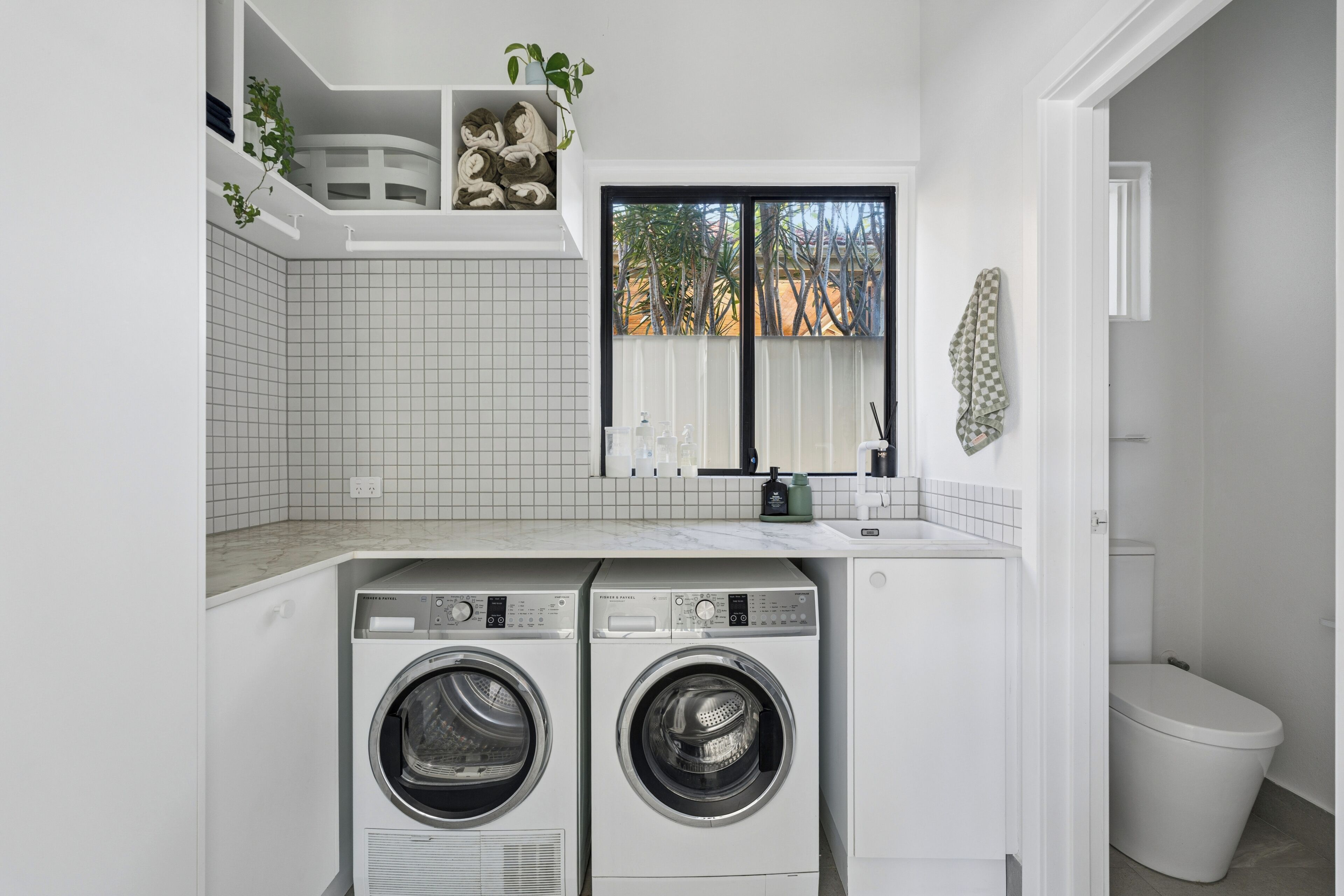 A laundry room with two front-loading washers, a tiled backsplash, a window with a view, and a white toilet.