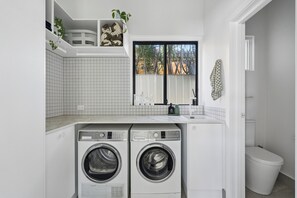 A laundry room with two front-loading washers, a tiled backsplash, a window with a view, and a white toilet. - Stunning Perth Summer Home with Pool, Gardens and Outdoor Living (Mount Hawthorn)