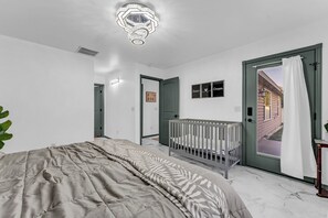 A bedroom with a bed, a crib, a door leading outside, and a wall-mounted light fixture.