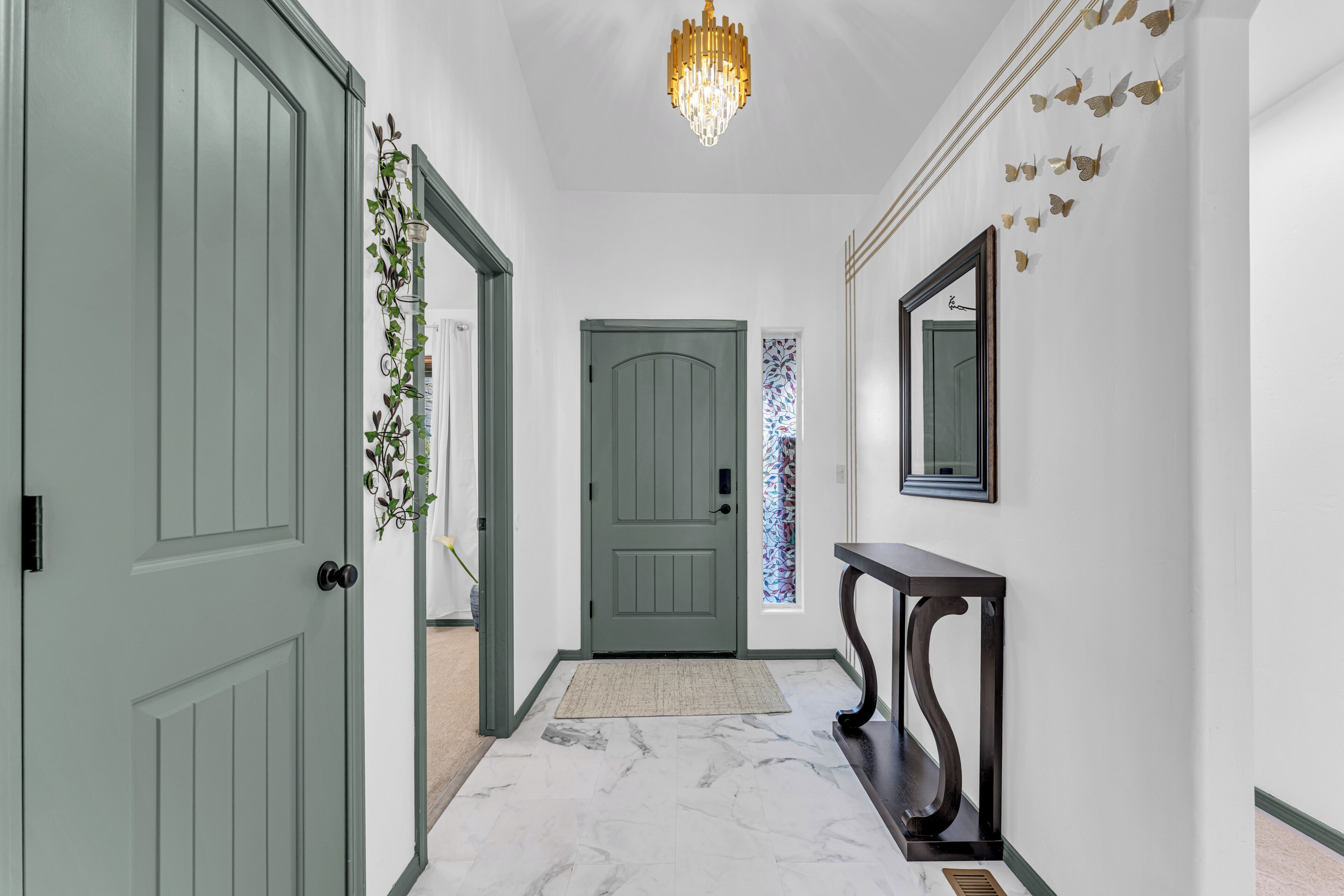 Un couloir avec une porte verte, un miroir fixé au mur, une plante décorative et un lustre.