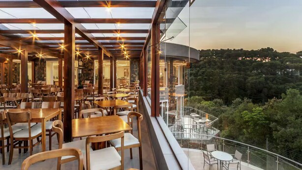 A restaurant with multiple dining tables and chairs, a glass railing overlooking a forested area, and a view of the surrounding landscape.